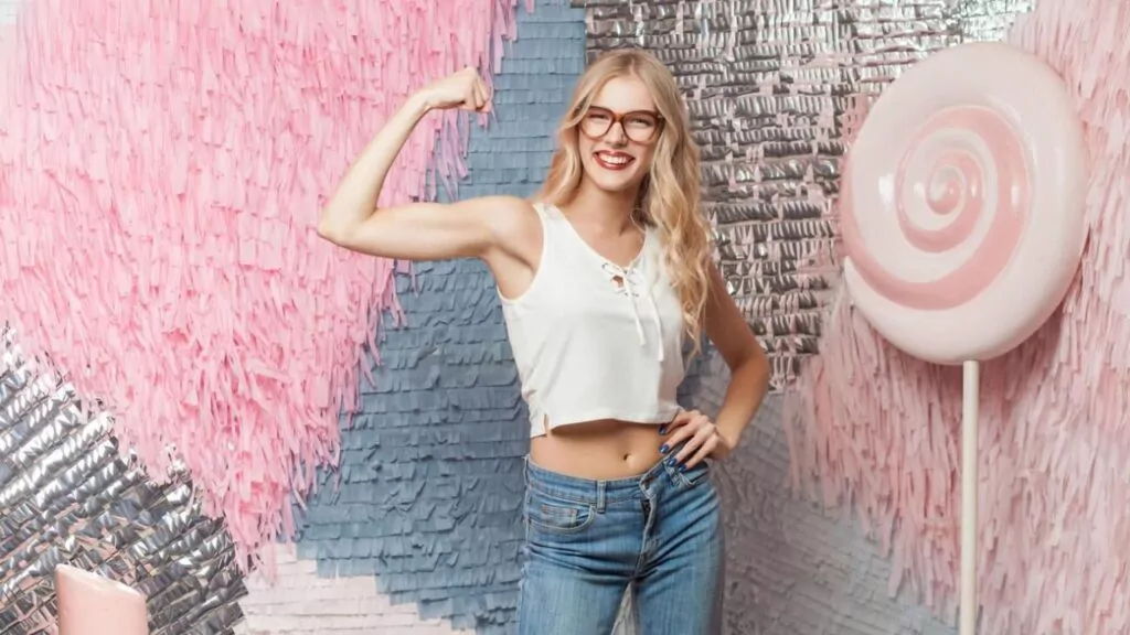 Funny young blonde girl with long hair in red glasses shows her muscles on colorful abstract background with huge candy, dressed in white top and jean