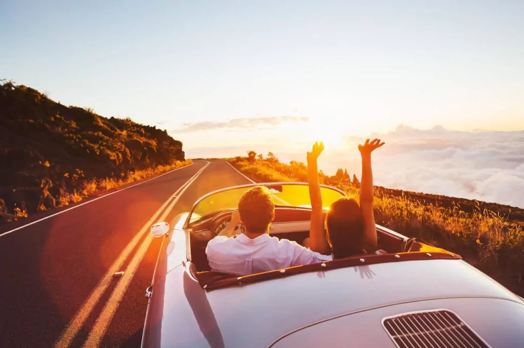 A happy couple is driving on road trips into the sunset in a classic vintage sports car.